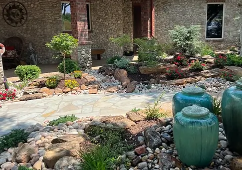 A landscaped courtyard featuring a stone path, vibrant garden beds, and three bubbling teal ceramic fountains.