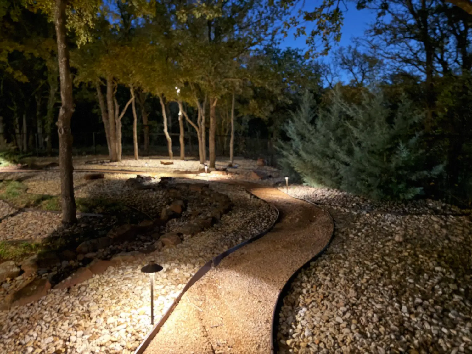 Nighttime luxury landscape design featuring illuminated pathways through mature trees and manicured shrubs